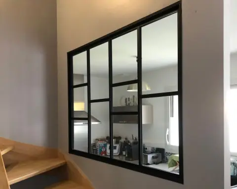 Une verrière noire posée dans une cloison, au-dessus d’un escalier en bois
