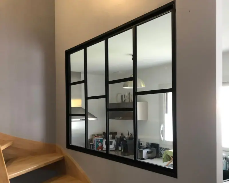 Une verrière noire posée dans une cloison, au-dessus d’un escalier en bois